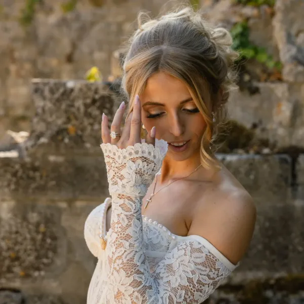Mariée souriante au chignon bouclé, gants de dentelle, devant un mur de pierres